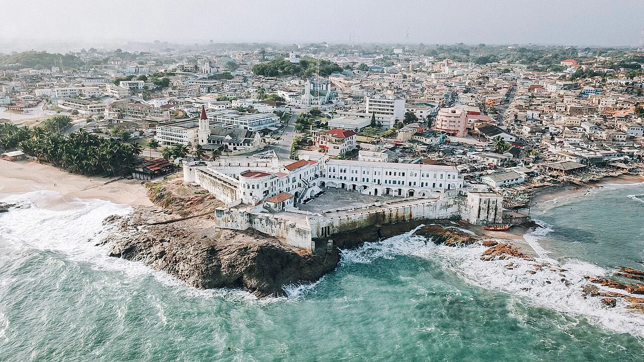 Aerial view of Cape Coast Castle above turquoise Atlantic waves, with the city behind it